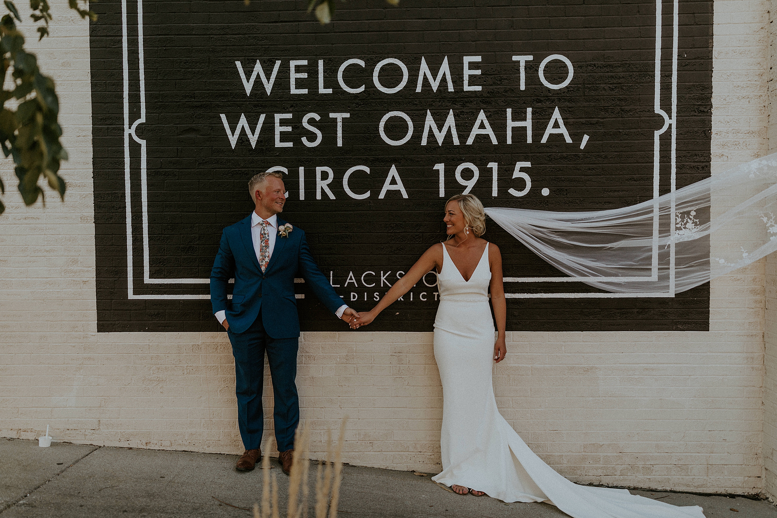 Bride and groom smiling at each other and holding hands as they pose in front of a street sign in Midtown Omaha for their wedding at the Omar