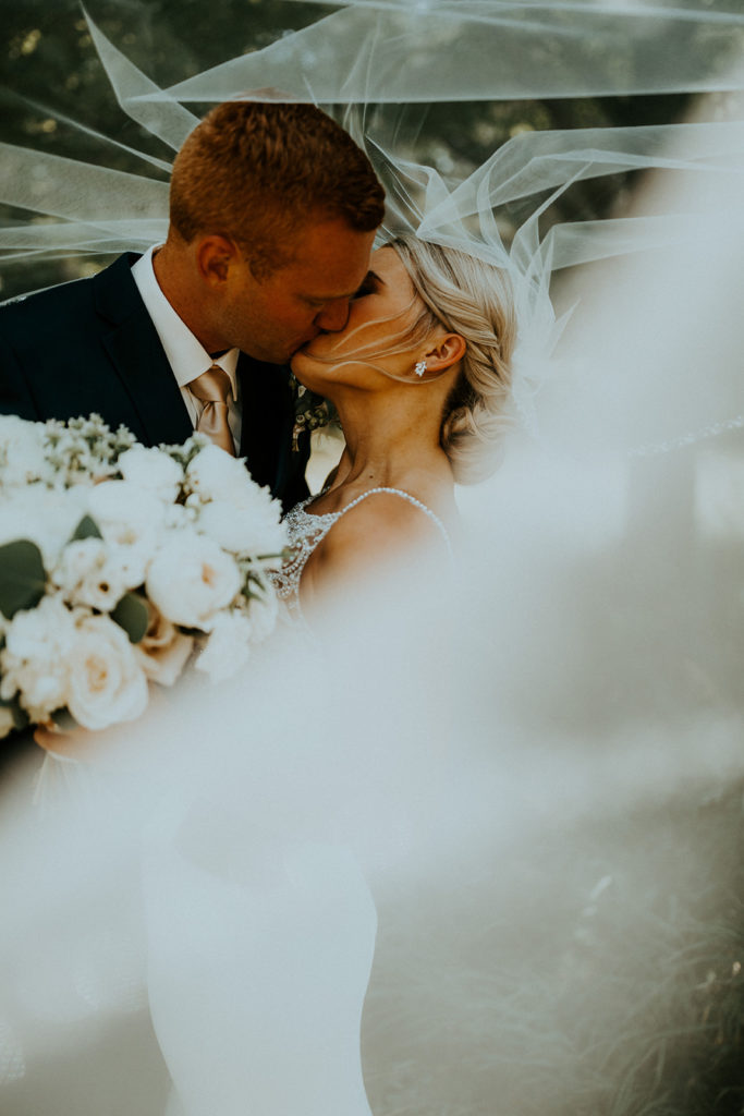 Newlywed couple share a kiss as bride's sheer veil flows in the wind, taken by McKenna Christine Photography