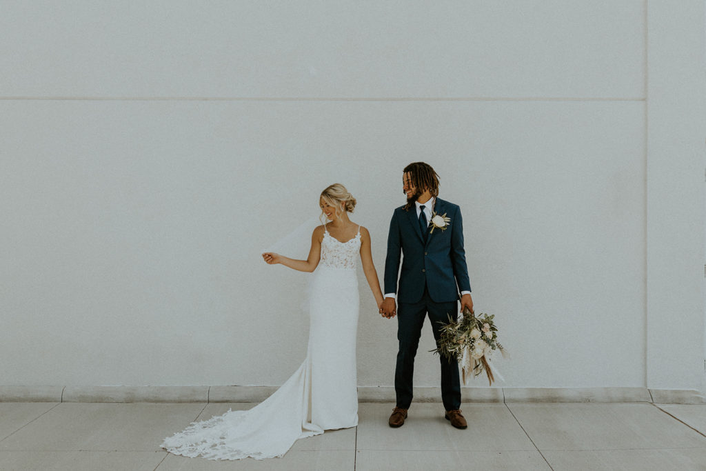 Bride and groom smiling and holding hands as they pose in front of white wall, shot by wedding photographer McKenna Christine