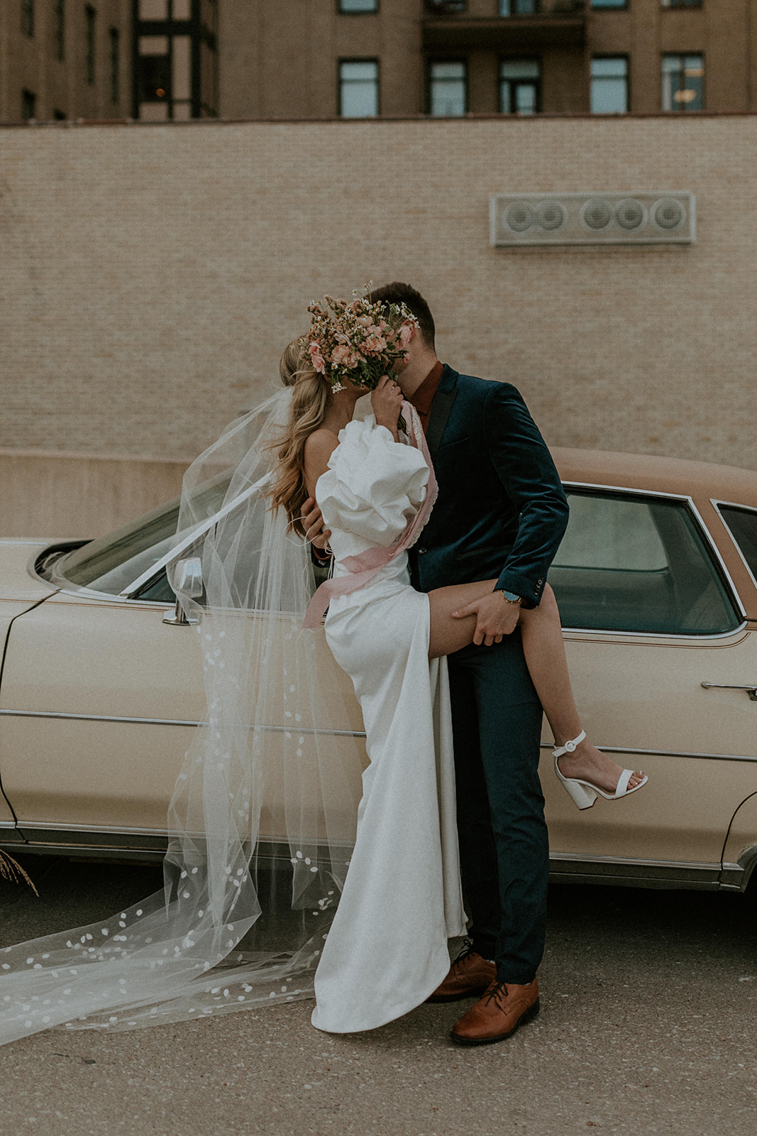 Couple pose in front of car with bride covering their faces with her bouquet, captured by McKenna Christine Photography
