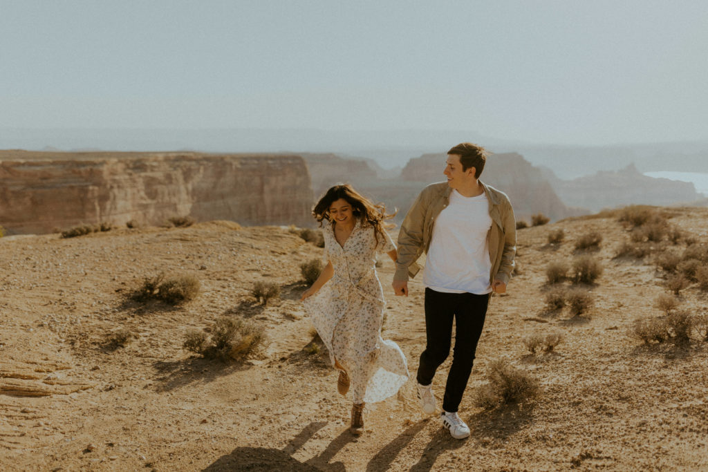 What to Wear For Engagement Photos + What NOT to Wear. Engaged couple holding hands as they run together with amazing view behind them.